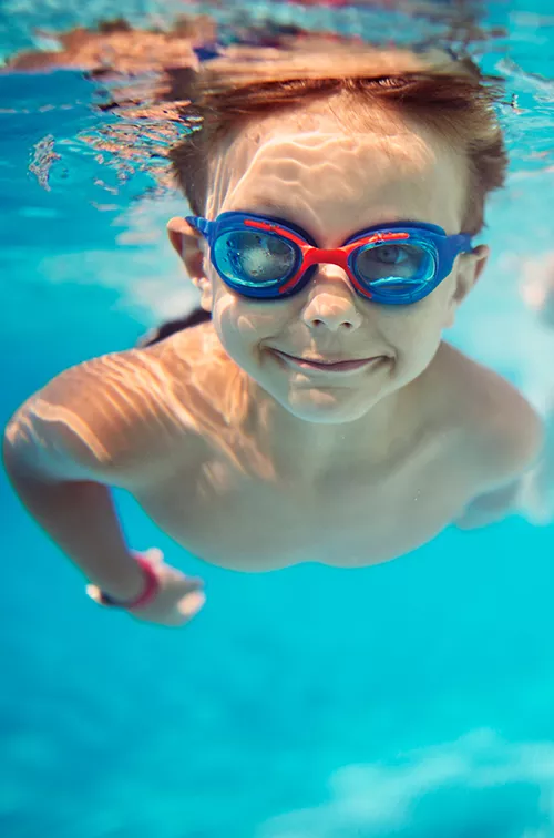 A young girl and boy swimming underwater in a pool while wearing goggles.