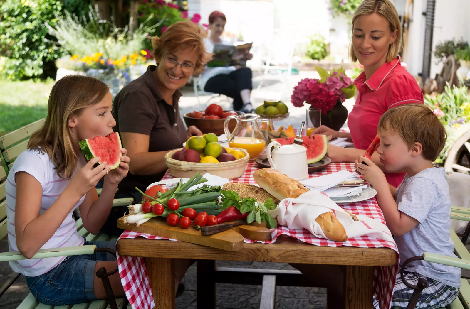 A family of four enjoys a summer picnic, outdoors, sitting at a picnic table enjoying summer foods.