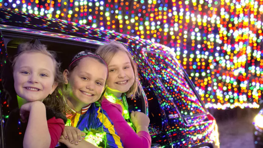 Three little girls smiling from their car while Christmas light illuminate around them