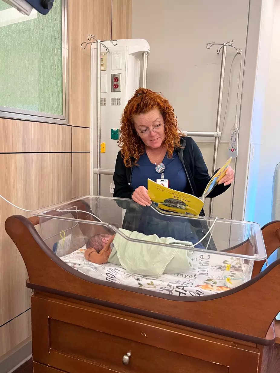 April Nelson, a nurse at AdventHealth for Children, stands over a crib, reading to a swaddled baby in the NICU.